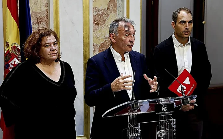 Tres políticos españoles compareciendo en rueda de prensa en el Congreso de los Diputados, con Enrique Santiago en el centro hablando ante un atril transparente y banderas de España y de la Unión Europea al fondo.