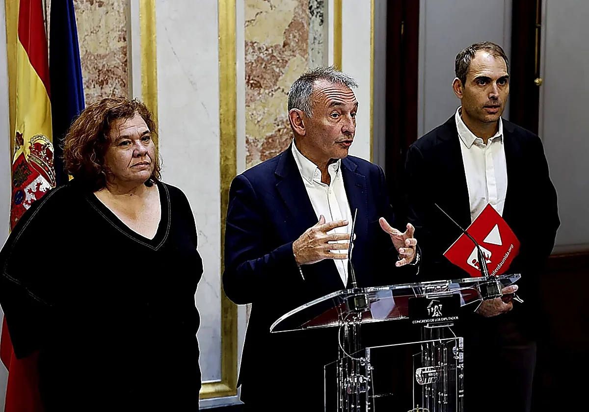 Tres políticos españoles compareciendo en rueda de prensa en el Congreso de los Diputados, con Enrique Santiago en el centro hablando ante un atril transparente y banderas de España y de la Unión Europea al fondo.