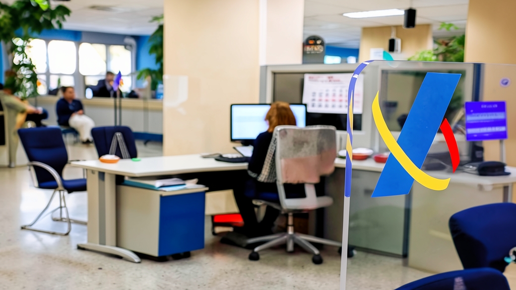 Oficina de la Agencia Tributaria con un logotipo azul y amarillo en primer plano sobre un separador de cristal transparente y empleados trabajando al fondo.