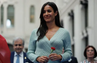 Fotografía de Begoña Villacís sonriendo sosteniendo un clavel rojo con personas al fondo.