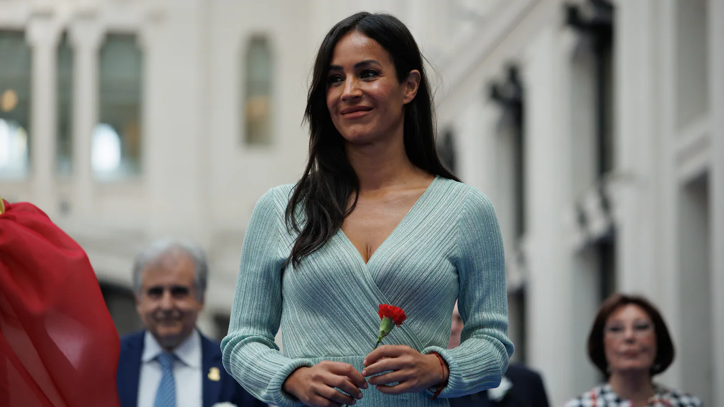 Fotografía de Begoña Villacís sonriendo sosteniendo un clavel rojo con personas al fondo.