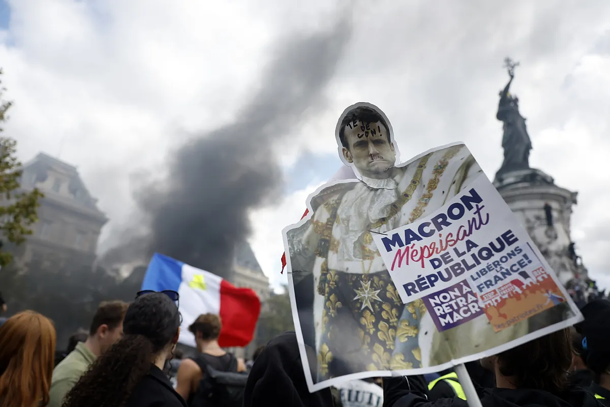 Protestas en Francia con manifestantes portando una pancarta crítica con Emmanuel Macron y una columna de humo negro de fondo en la calle.