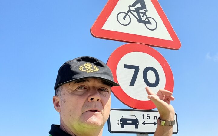 Miembro de la Guardia Civil de tráfico señalando un cartel de señalización vial con límite de velocidad y advertencia de ciclistas bajo un cielo azul despejado.