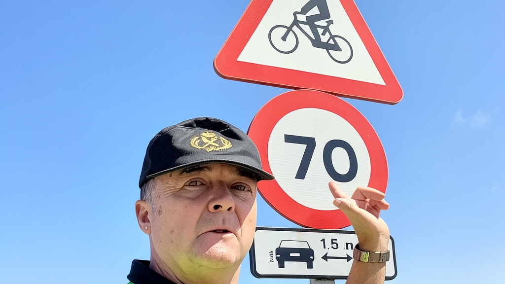 Miembro de la Guardia Civil de tráfico señalando un cartel de señalización vial con límite de velocidad y advertencia de ciclistas bajo un cielo azul despejado.