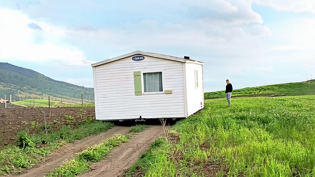 Fotografía editorial de una casa móvil blanca instalada en una parcela rústica con un camino de tierra y una persona de pie en el campo verde.