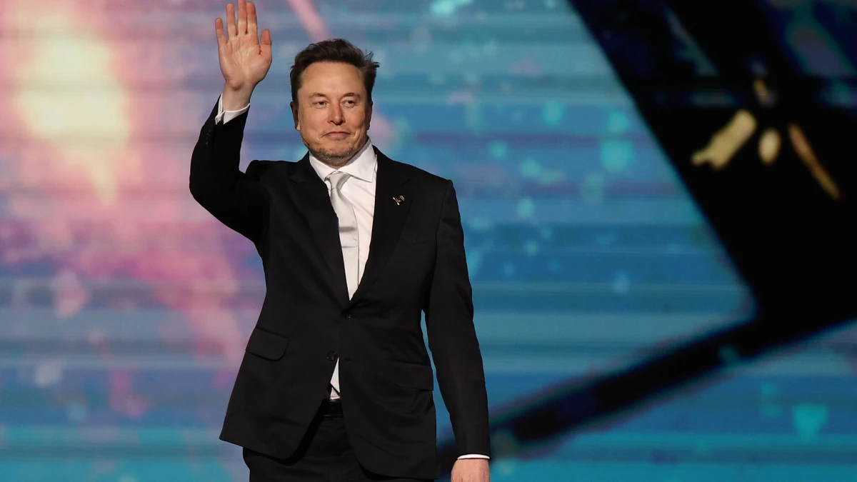 Fotografía editorial de Elon Musk saludando con la mano levantada frente a un fondo azulado en un evento corporativo.