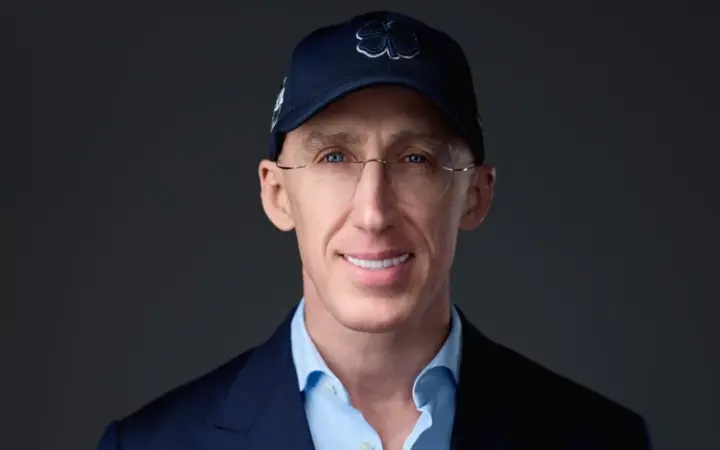 Retrato de Uri Poliavich con gorra azul, gafas y americana oscura sobre fondo neutro.