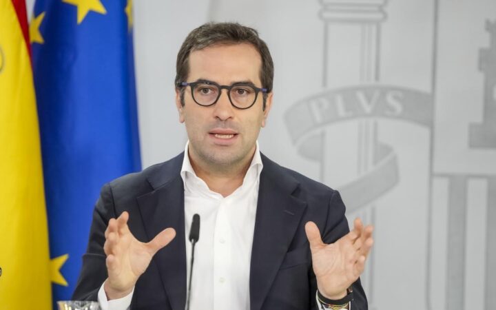 Fotografía institucional de un político con traje, camisa blanca y gafas, gesticulando con las manos durante una rueda de prensa en España.