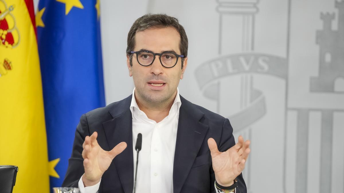 Fotografía institucional de un político con traje, camisa blanca y gafas, gesticulando con las manos durante una rueda de prensa en España.
