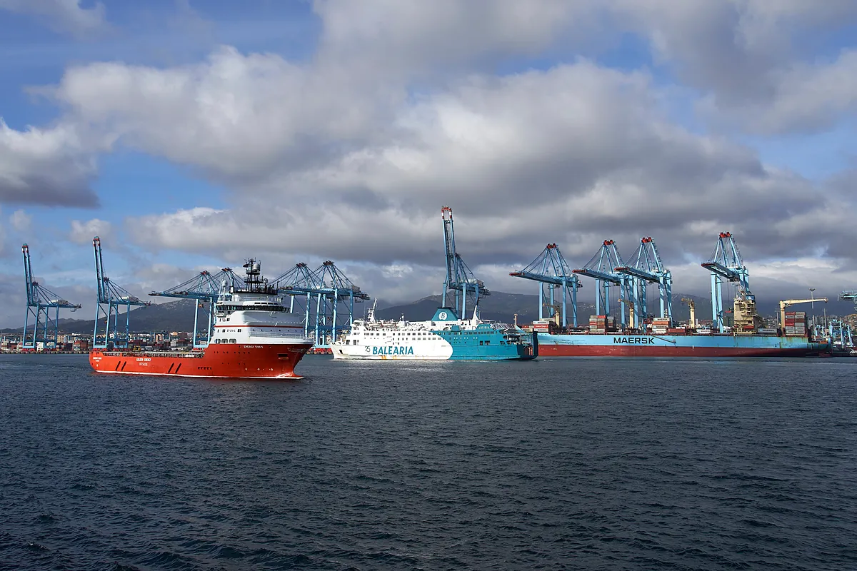 Vista panorámica de un puerto marítimo con barcos de carga, un ferri y grúas portuarias bajo un cielo nublado.