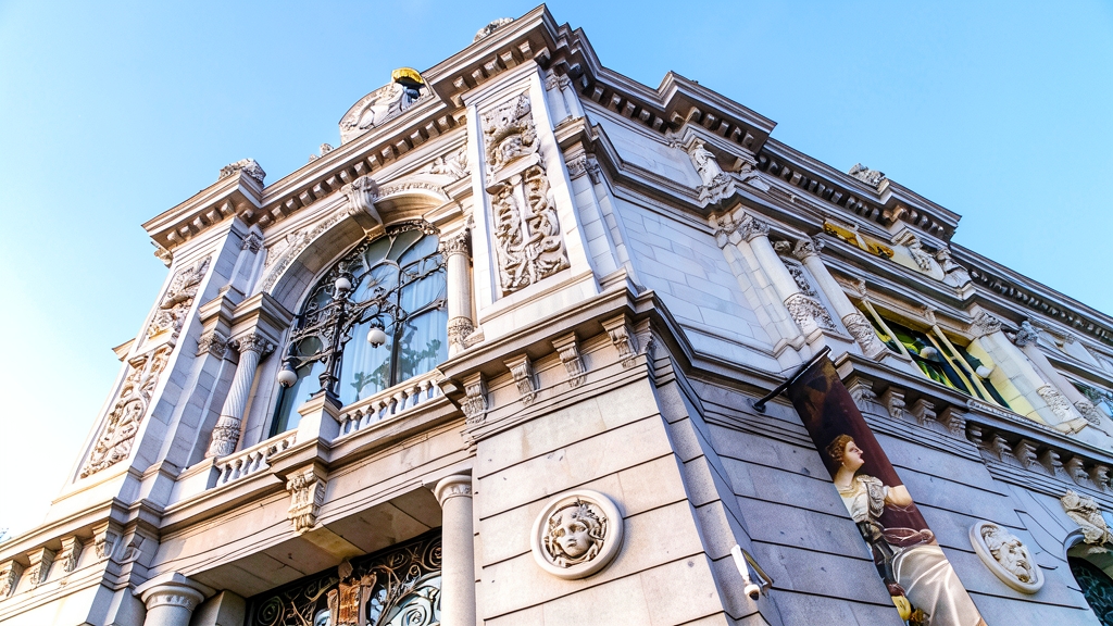 Fotografía en contrapicado de la fachada principal del edificio del Banco de España, mostrando su ornamentada arquitectura clásica con esculturas, relieves y un gran ventanal con reja de hierro forjado.