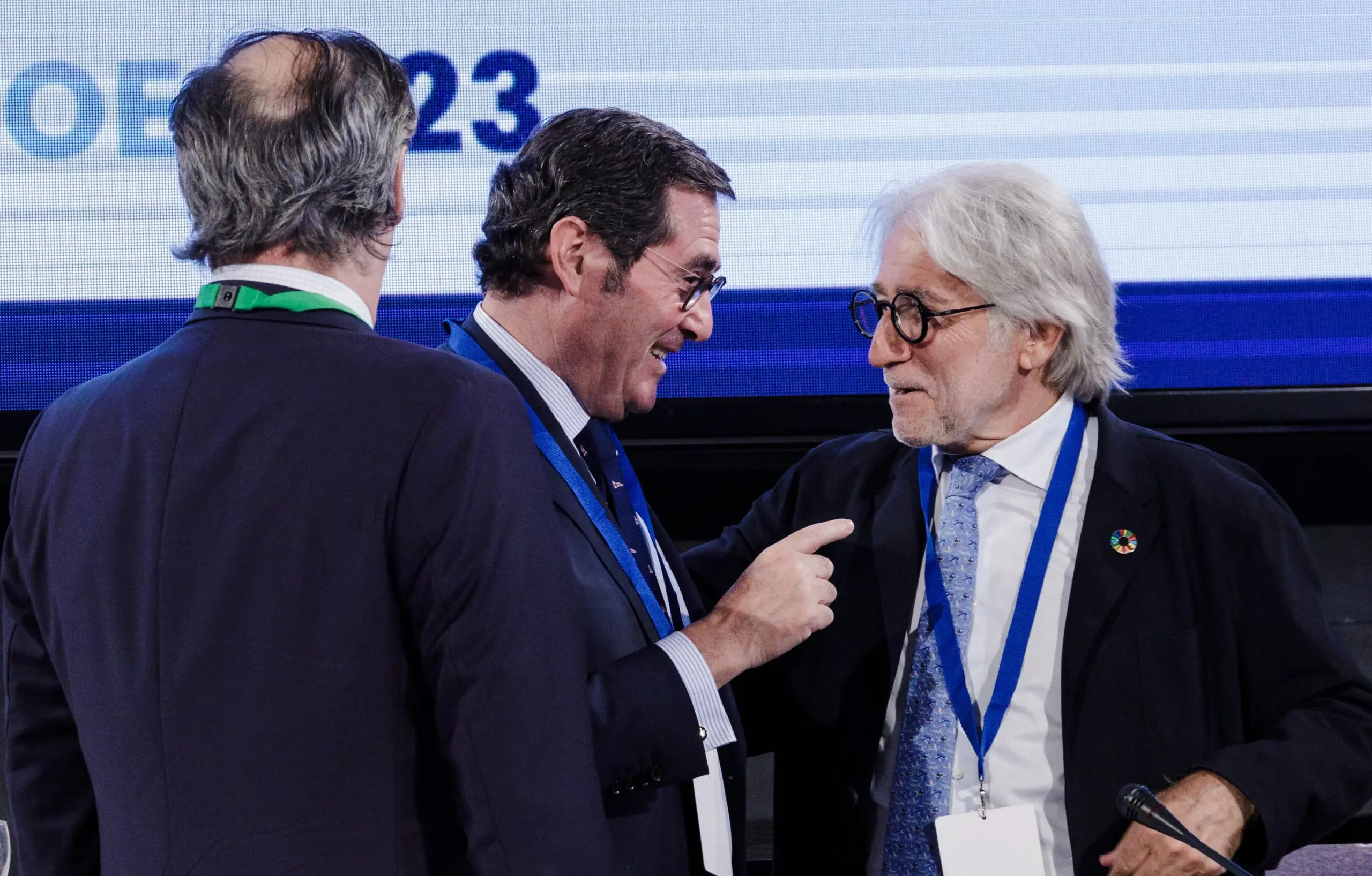 Tres hombres de negocios en un evento corporativo charlando, con pantallas de fondo y micrófonos.