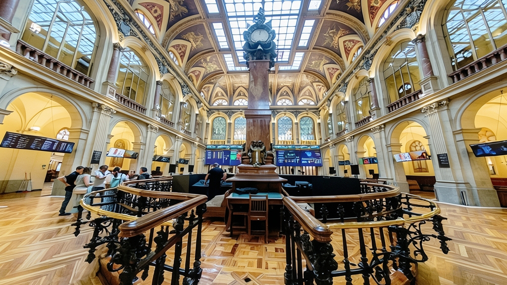 Fotografía gran angular del interior de la Bolsa de Madrid con pantallas de cotización, columnas ornamentadas y una barandilla de madera en primer plano.