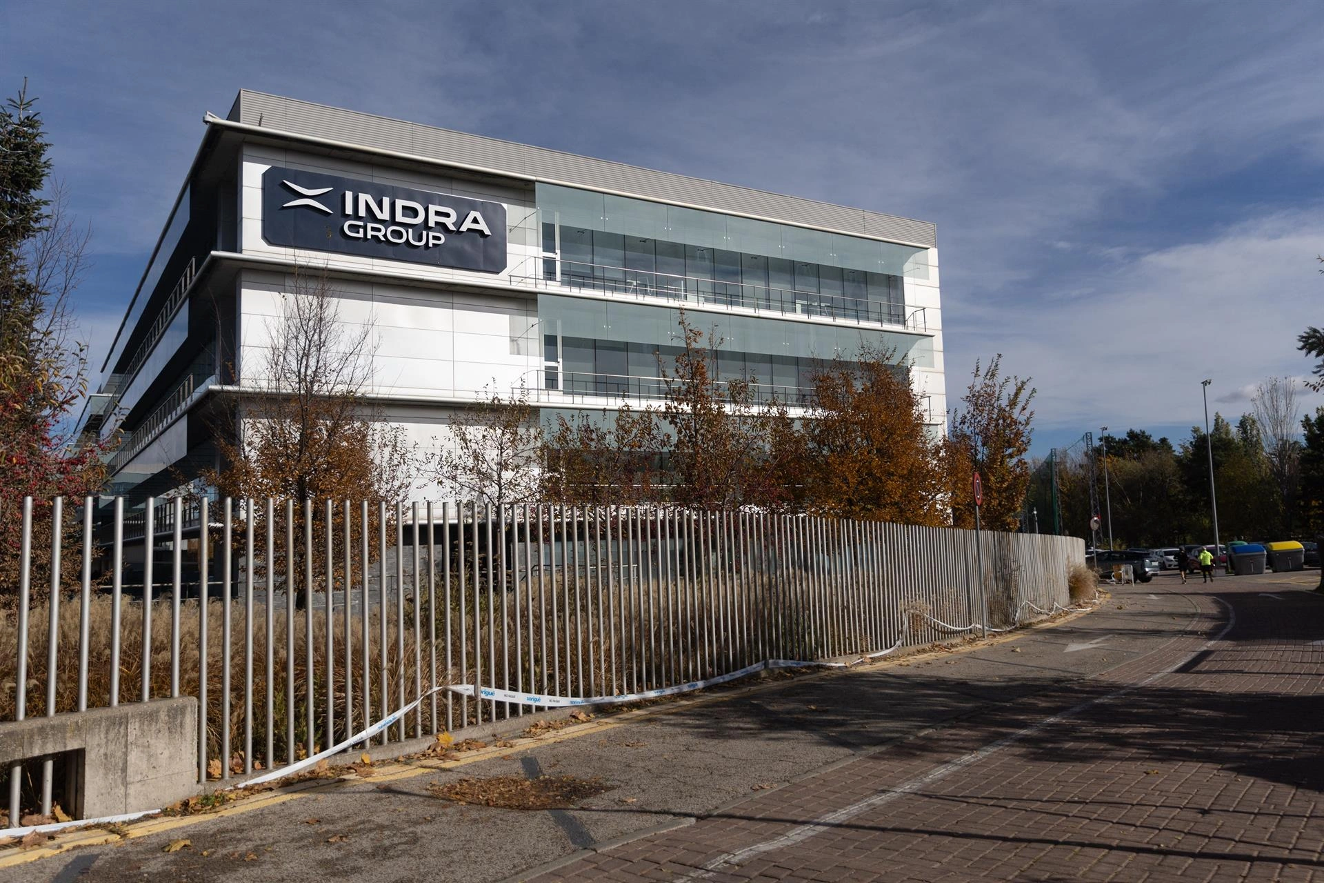 Sede corporativa del grupo Indra Group bajo un cielo parcialmente nublado, con el logotipo visible en la fachada de un edificio moderno de oficinas acristalado y protegido por una valla metálica exterior.