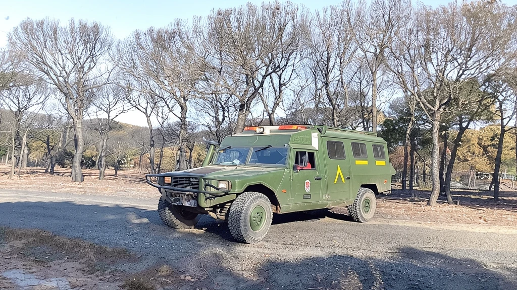 Vehículo táctico militar verde estacionado en un camino de tierra junto a un bosque con árboles quemados.