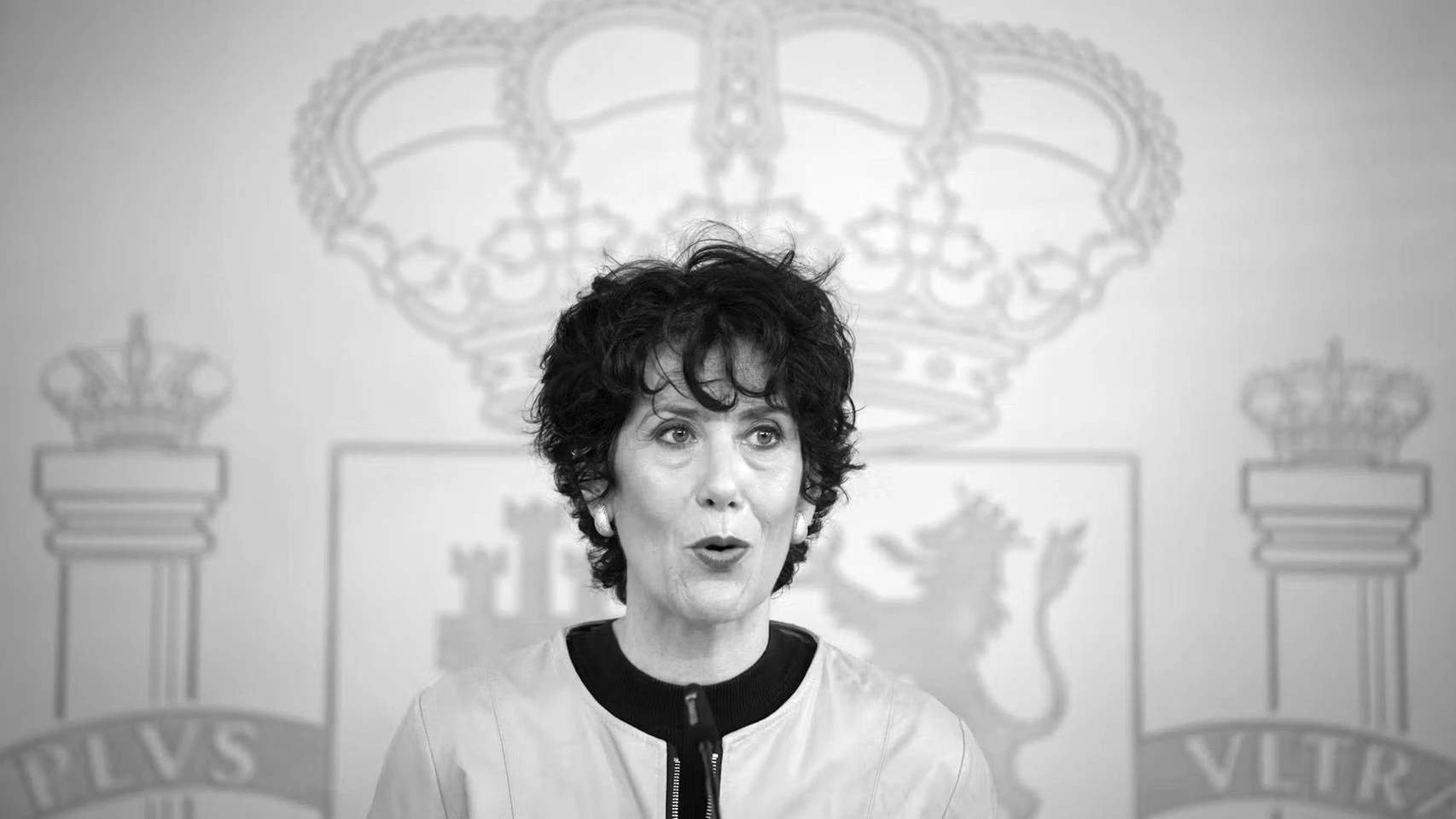 Fotografía en blanco y negro de Elma Saiz compareciendo en rueda de prensa ante el escudo de España.