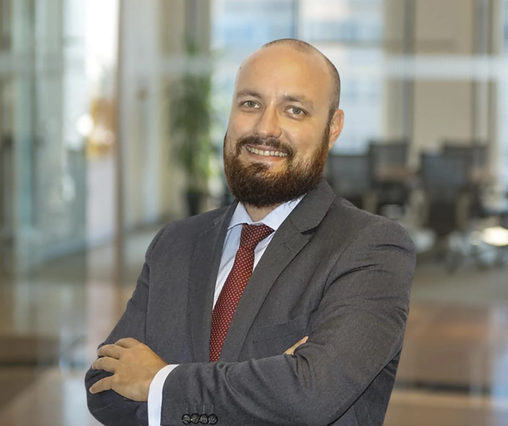 Retrato corporativo de un hombre calvo con barba, sonriendo, vestido con traje gris y corbata roja, con los brazos cruzados en una oficina.