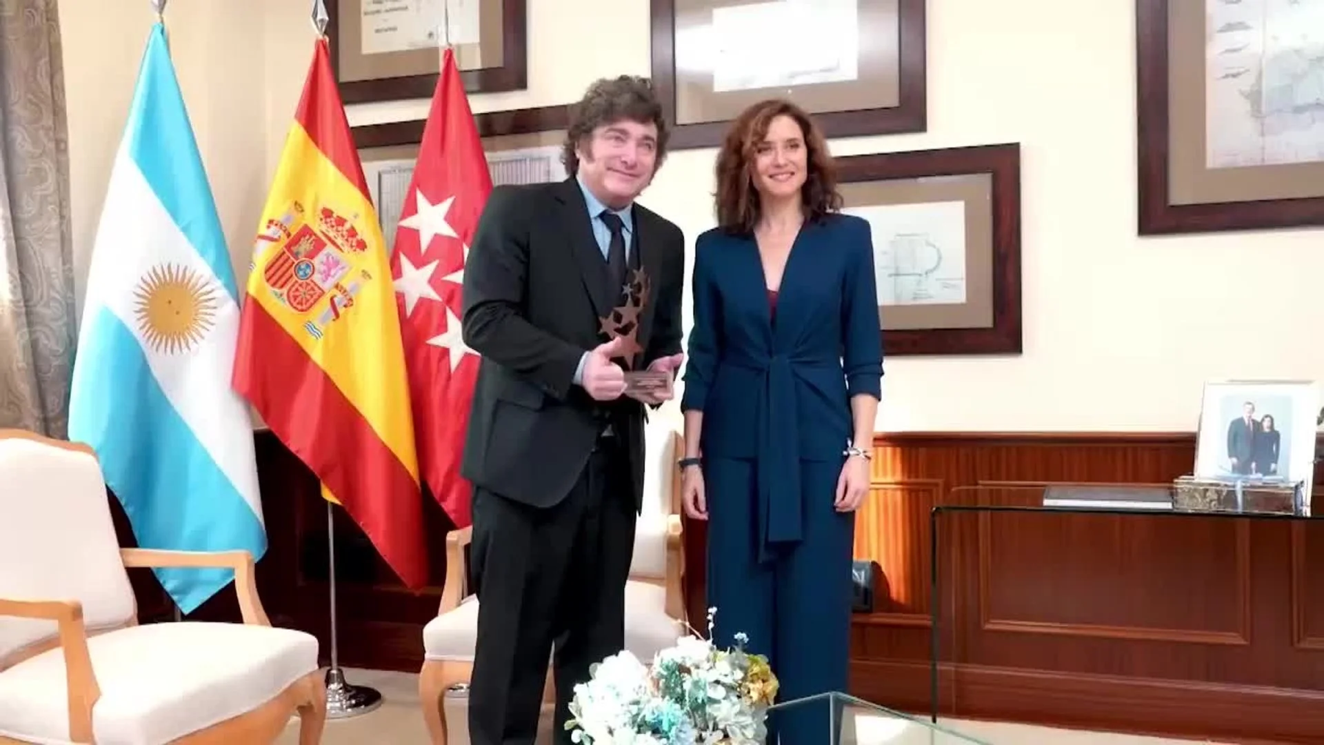 Javier Milei e Isabel Díaz Ayuso posan juntos con una estatuilla de estrellas y las banderas de Argentina, España y la Comunidad de Madrid al fondo.
