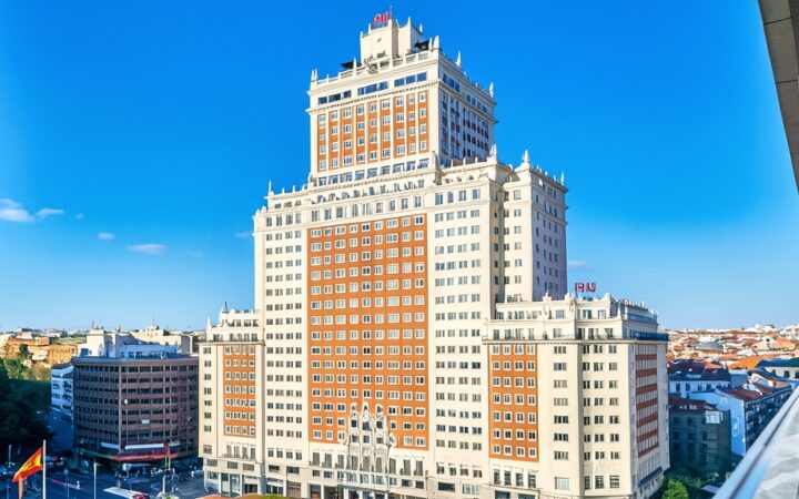 Fotografía del edificio del Hotel Riu Plaza España en Madrid bajo un cielo azul despejado, con la bandera de España abajo a la izquierda.