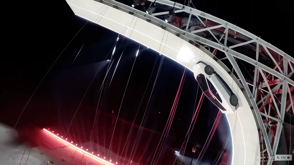 Fotografía nocturna en ángulo picado que muestra un coche deportivo blanco sobre una rampa curva iluminada por luces rojas y blancas, con estructuras metálicas de soporte a un lado.
