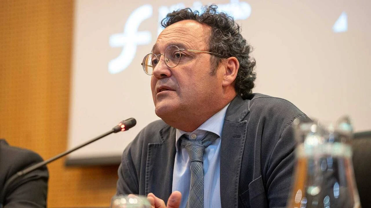 Fotografía editorial de Álvaro García Ortiz hablando en una mesa con micrófono y proyector al fondo.