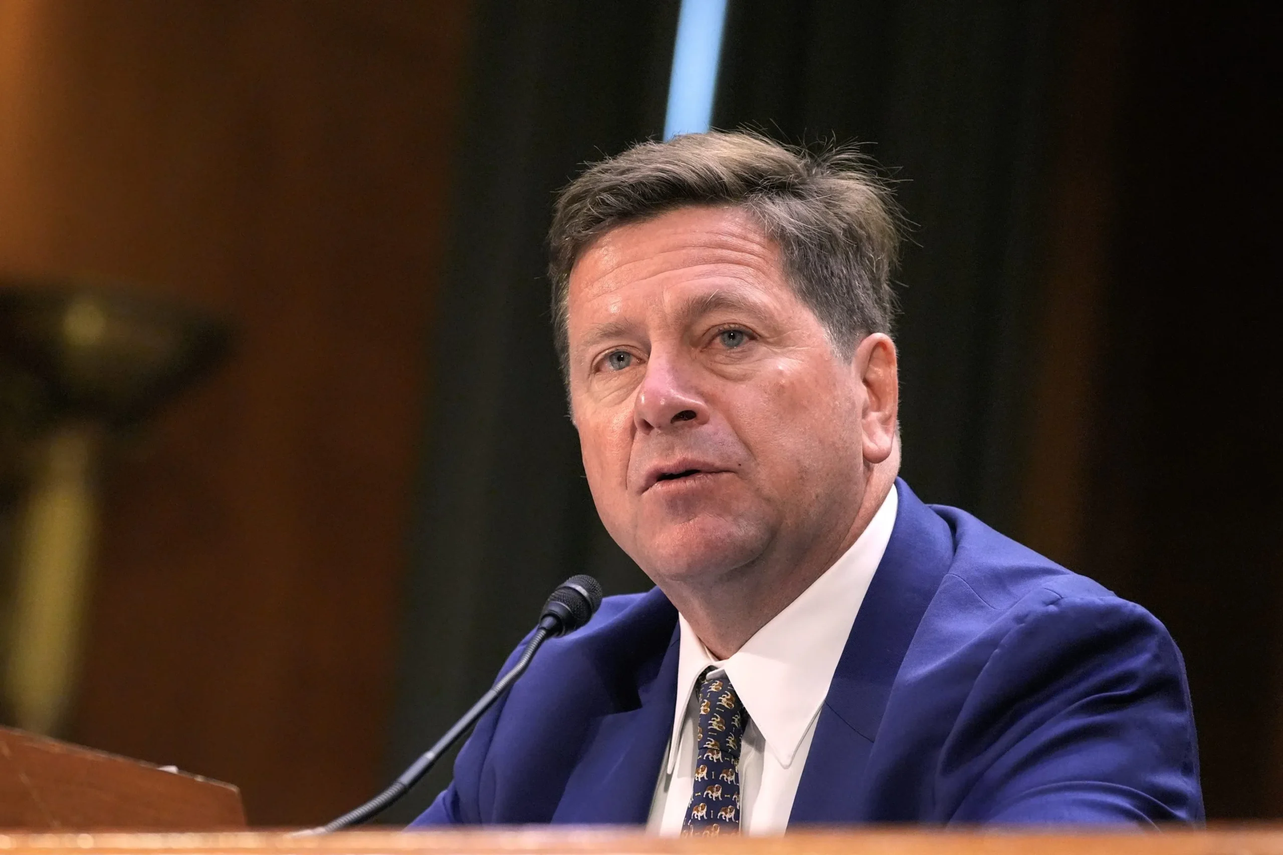 Fotografía editorial de Jay Clayton hablando en una audiencia pública ante un micrófono.
